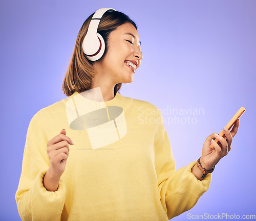 Image of Music, phone and happy Asian woman in studio for social media, internet meme and online chat. Headphones, smile and female person on smartphone listening to audio, song and track on purple background