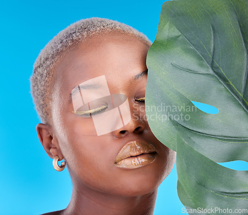 Image of Makeup, plant and a black woman on a blue background for a wellness aesthetic. Face, beauty and an African girl or model with a leaf and organic cosmetics isolated on a studio backdrop for health