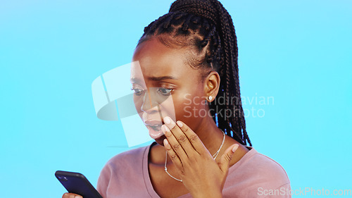 Image of Phone, wow and surprise with a black woman in studio on a blue background looking shocked at news. Mobile, social media or shock and a female indoor with an omg or wtf hand gesture over her mouth