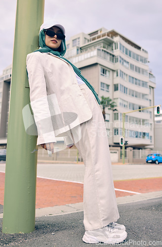 Image of Portrait, city fashion or religion with a muslim woman leaning against a pole in a cap and scarf for contemporary style. Islam, faith or hijab with a trendy young arab person outside in an urban town