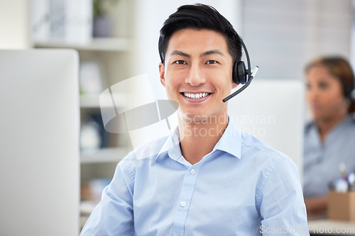 Image of Portrait of asian call centre agent talking on headset while working on computer in office. Confident and smiling businessman consulting and operating a helpdesk for customer sales and service suppor