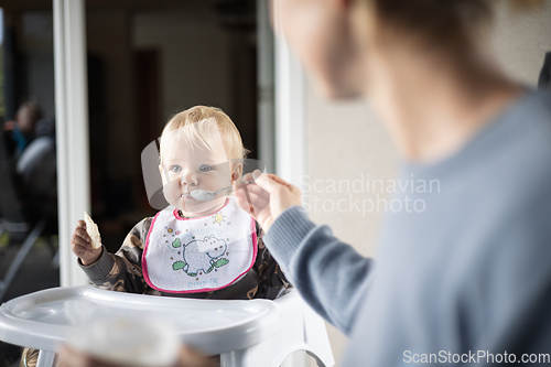 Image of Mother spoon feeding her baby boy child in baby high chair with fruit puree at dinning table at home. Baby solid food introduction concept.