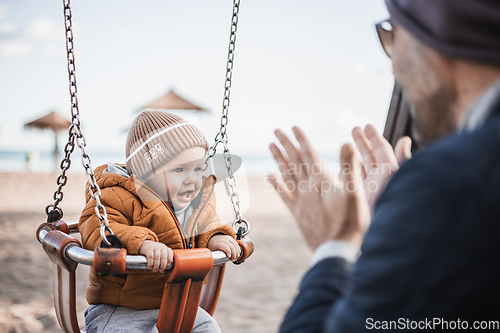 Image of Father pushing hir cheerful infant baby boy child on a swing on sandy beach playground outdoors on nice sunny cold winter day in Malaga, Spain.