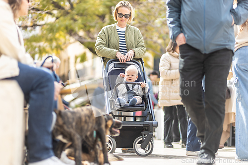 Image of Mother walking and pushing his infant baby boy child in stroller in crowd of people wisiting sunday flea market in Malaga, Spain.