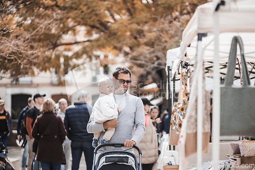 Image of Father walking carrying his infant baby boy child and pushing stroller in crowd of people wisiting sunday flea market in Malaga, Spain