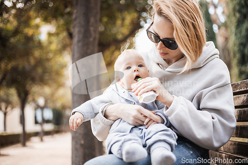 Image of Mother sitting on bench in urban park, holding her infant baby boy child in her lap and feeding him baby food on nice spring sunny day
