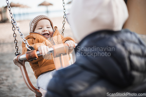 Image of Mother pushing her cheerful infant baby boy child on a swing on sandy beach playground outdoors on nice sunny cold winter day in Malaga, Spain.