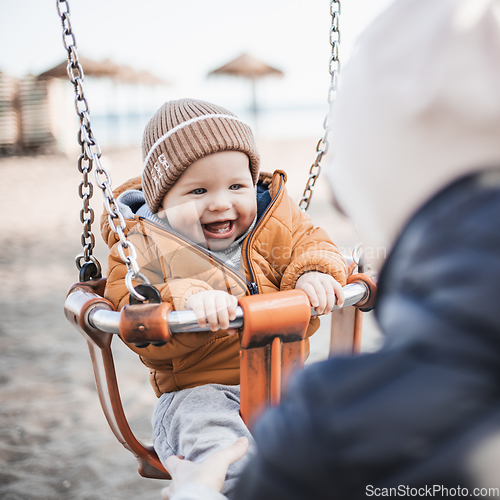 Image of Mother pushing her cheerful infant baby boy child on a swing on sandy beach playground outdoors on nice sunny cold winter day in Malaga, Spain.