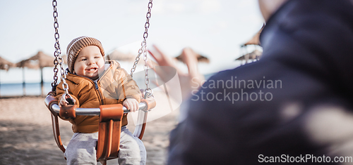 Image of Mother pushing her cheerful infant baby boy child on a swing on sandy beach playground outdoors on nice sunny cold winter day in Malaga, Spain.