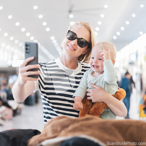 Image of Mother taking selfie with mobile phone, while traveling with child, holding his infant baby boy at airport, waiting to board a plane. Travel with kids concept.