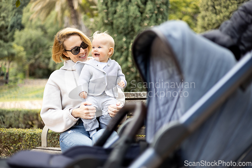 Image of Mother sitting on bench in urban park, laughing cheerfully, holding her smiling infant baby boy child in her lap having baby stroller parked by their site.