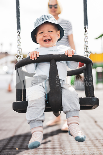 Image of Mother pushing her infant baby boy child on a swing on playground outdoors.