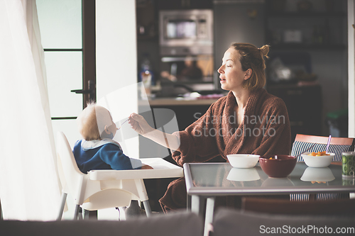 Image of Cheerful mother wearing bathrope spoon feeding her infant baby boy child sitting in high chair at the dining table in kitchen at home in the morning.