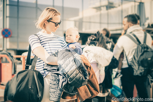 Image of Motherat travelling with his infant baby boy child. Mom holding travel bag and her infant baby boy child while queuing for bus in front of airport terminal station.