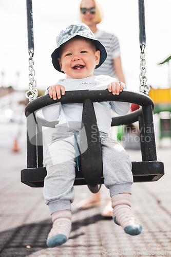 Image of Mother pushing her infant baby boy child on a swing on playground outdoors.