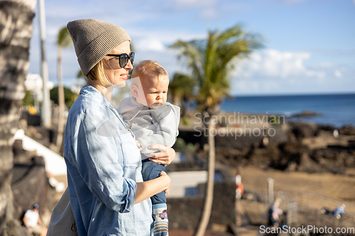 Image of Fashinable mother holding her infant boy child in lap enjoing panoramic view along palm lined beach promenade of Puerto del Carmen, Lanzarote, Canary islands, Spain.