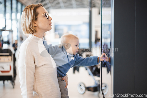 Image of Mother traveling with child, holding his infant baby boy at airport terminal, checking flight schedule, waiting to board a plane. Travel with kids concept.