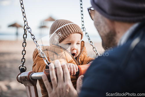 Image of Father pushing hir cheerful infant baby boy child on a swing on sandy beach playground outdoors on nice sunny cold winter day in Malaga, Spain.