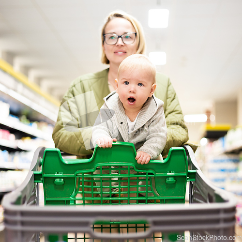 Image of Mother pushing shopping cart with her infant baby boy child down department aisle in supermarket grocery store. Shopping with kids concept.