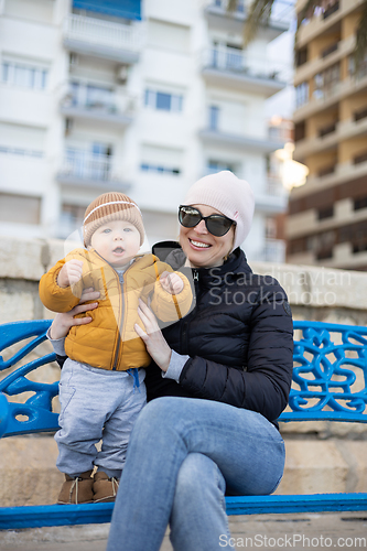 Image of Young mother with her cute infant baby boy child on bench in city park.