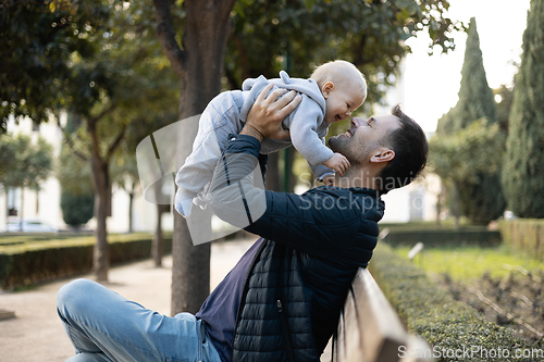 Image of Father holding and lifting his cute infant baby boy child sitting on wooden bench in urban city park. Dad and son enjoing a pristine moment of happiness, smiling and laughing