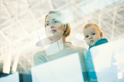 Image of Thoughtful young mother looking trough window holding his infant baby boy child while waiting to board an airplane at airport terminal departure gates. Travel with baby concept.