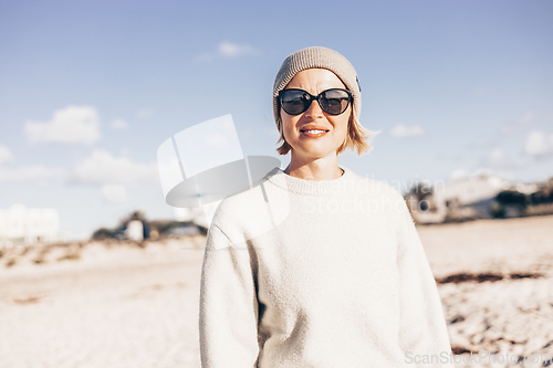 Image of Portrait of young stylish woman wearing wool sweater, wool cap and sunglasses on long sandy beach in spring