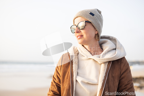 Image of Portrait of young stylish woman wearing brown padded jacket, hoodie, wool cap and sunglasses on long sandy beach in spring.