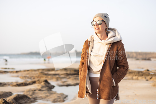 Image of Portrait of young stylish woman wearing brown padded jacket, hoodie, wool cap and sunglasses on long sandy beach in spring.