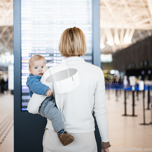 Image of Mother traveling with child, holding his infant baby boy at airport terminal, checking flight schedule, waiting to board a plane. Travel with kids concept.