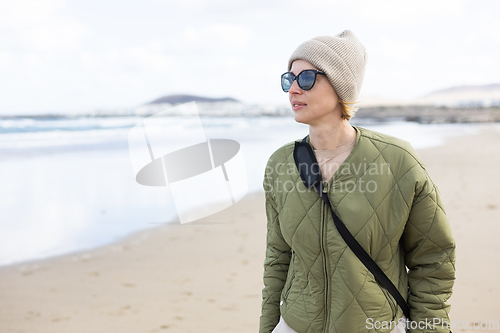 Image of Portrait of young stylish woman wearing green padded jacket, hoodie, wool cap and sunglasses on long sandy beach in spring.