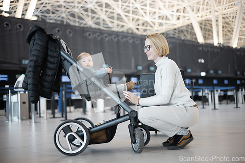 Image of Motherat interacting with her infant baby boy child in stroller while travelling at airport terminal station. Travel with child concept.