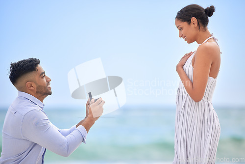 Image of Engagement, proposal and romance with a couple on the beach for a milestone on a blue sky background. Love, summer or nature with a man down on one knee to ask his girlfriend the marriage question