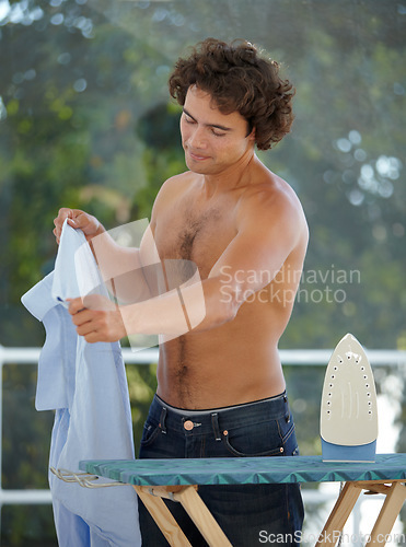 Image of Iron, shirt and a topless man doing laundry outdoor on the deck of his home for housework during the day. Clothes and ironing board with a handsome young male person doing household chores