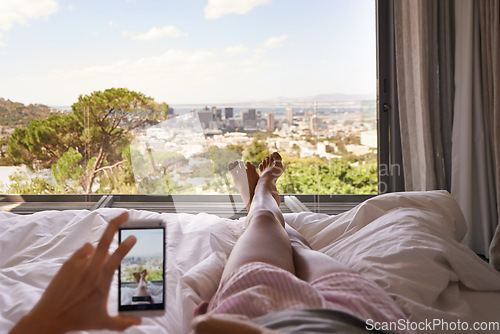 Image of Bed, phone and window view with feet, relax and photography for blog, review and screen in hotel. Woman, hand and smartphone for profile picture with cityscape, nature or social media app in bedroom