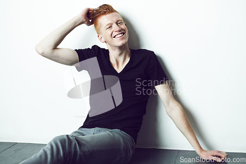 Image of Portrait, ginger and man sitting on a floor with hand on head or relax or smile for fashion and against white wall. Happy, unique style and excited male model with red hair laugh for fun in studio