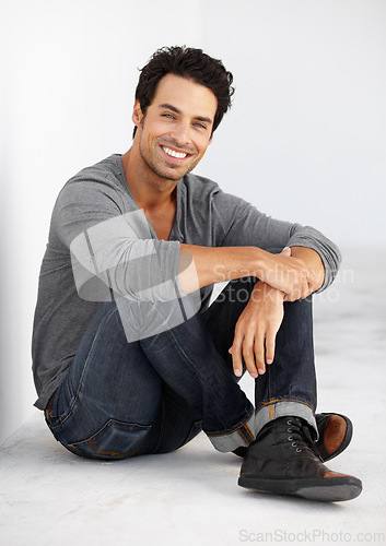 Image of Floor, portrait and happy man isolated on a wall background with casual clothes, confidence and relax. Handsome model or Portugal person smile and sitting on ground in minimal room with cool fashion