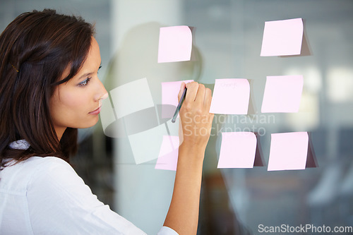 Image of Business woman, writing and planning for brainstorming, strategy or ideas on glass board at the office. Female employee working on tasks for reminder, sticky note or project plan at the workplace