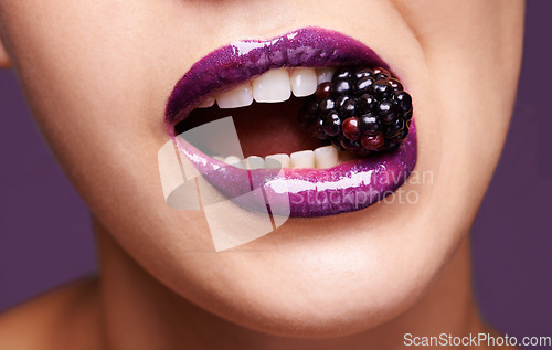 Image of Woman lips, purple lipstick with blackberry and makeup, shine and creativity with beauty isolated on studio background. Closeup of fruit between female model teeth, cosmetic product and cosmetology