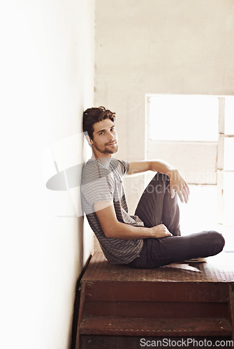 Image of Portrait, relax and stairs with a man sitting by a window on the landing of a staircase while alone in his home. Happy, content and lifestyle with a handsome young male person laid-back in a house