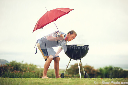 Image of Umbrella, rain and a man outdoor to barbecue food for cooking or insurance in the winter season. Storm, weather and grill with a male person getting wet while trying to bbq on a grass lawn or garden