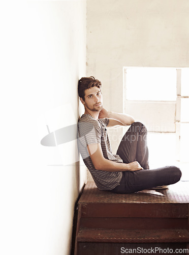 Image of Portrait, depression and alone with a man on a staircase in his home, sitting on the landing by a window. Lonely, mental health and relax with a handsome young male on steps or stairs in his house