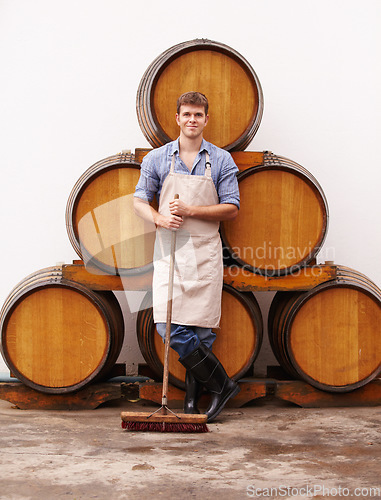 Image of Portrait, broom and oak barrels with a man cleaning the wine cellar of a beverage distillery. Confident, apron and a male cleaner in a winery for the production, fermentation or storage of alcohol