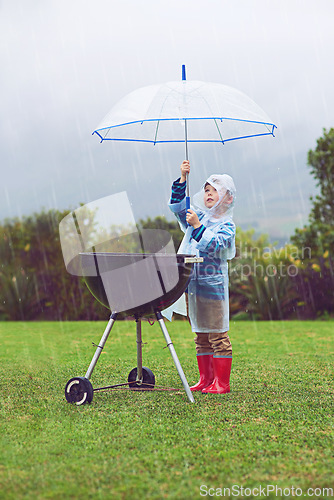 Image of Rain, umbrella and a boy covering a barbecue outdoor on a field while cooking food alone in winter. Raincoat, kids and insurance with a young male child trying to protect a bbq during stormy weather