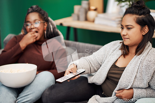 Image of Girl friends, watching tv and remote in home living room on a couch with snack eating. Series, movie and women together in a lounge with a female friend looking at a television in house with popcorn