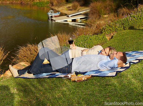 Image of Couple, picnic and lying by lake with wine outdoor, having fun and bonding on vacation. Portrait, alcohol and man and woman by river to relax, happiness and enjoying romantic time together in summer.