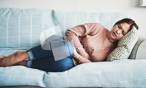 Image of Woman, stomach ache or pain on home sofa with menstrual or period cramps in lounge. Sick, abdomen or colon problem of a female person with hands on tummy for constipation, digestion or virus