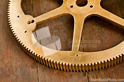 Image of Clock gear, gold and wheel cog on a wood table with golden machinery and gears. Metal, closeup and steel with machine detail and maintenance on wooden tabletop with brass parts and wheels on surface