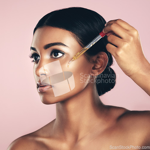 Image of Face, skincare serum and woman in studio isolated on a pink background. Dermatology, cosmetics and Indian model with hyaluronic acid, essential oil or vitamin c dropper for healthy skin and beauty.