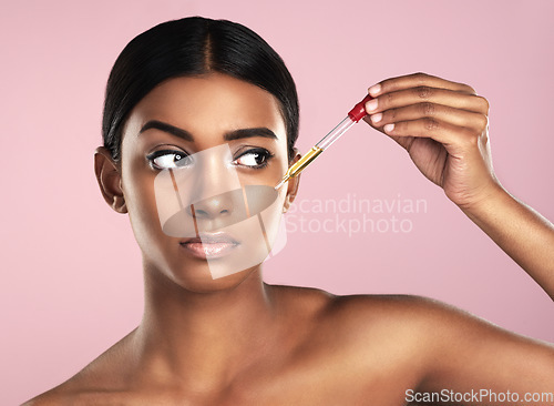 Image of Face, skincare serum and serious woman in studio isolated on a pink background. Dermatology, cosmetics and Indian model with hyaluronic acid, essential oil or apply vitamin c dropper for healthy skin